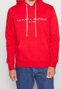 Sudadera roja con capucha, que cuenta con un bolsillo frontal, capucha ajustable con cordón y el logotipo bordado "TOMMY HILFIGER EST. 1985". Textura de tela suave.