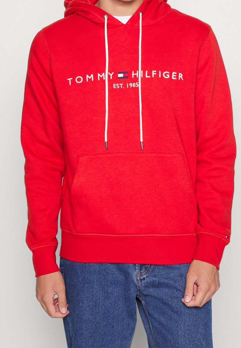 Sudadera roja con capucha, que cuenta con un bolsillo frontal, capucha ajustable con cordón y el logotipo bordado "TOMMY HILFIGER EST. 1985". Textura de tela suave.