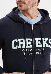 Felpa con zip colore navy con cordini, dotata della grafica bianca e azzurro chiaro "CREEKS ORIGINAL Dal 1974", realizzata in morbido tessuto.