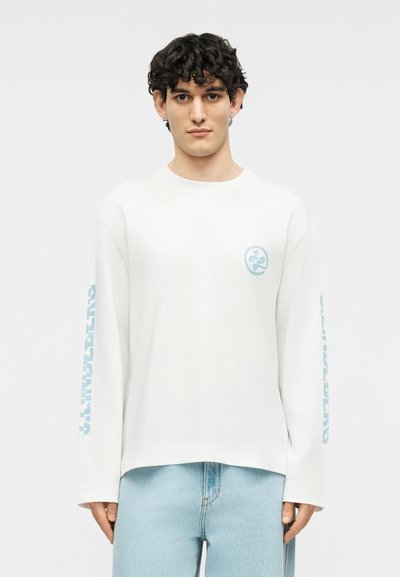 Νέος άνδρας με σγουρά σκούρα μαλλιά φοράει λευκό πουκάμισο μακρυμάνικο με μπλε κείμενο στα μανίκια και ανοιχτόχρωμο μπλε τζιν, στέκεται μπροστά σε απλό φόντο.