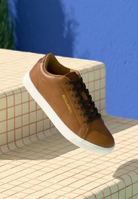 Bruine leren sneaker met een ronde neus, witte rubberen zool, contrasterende donkere veters en een gouden merkaanduiding aan de zijkant, tegen een geruit achtergrond.
