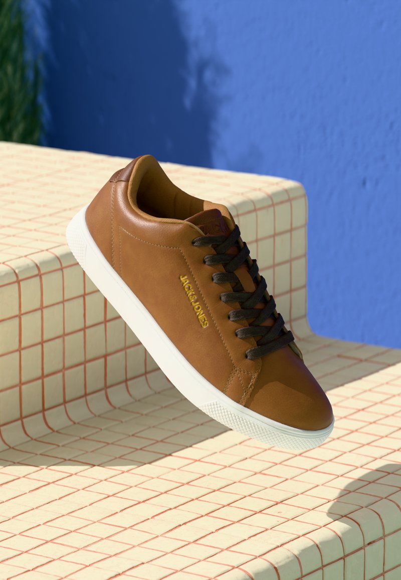 Bruine leren sneaker met een ronde neus, witte rubberen zool, contrasterende donkere veters en een gouden merkaanduiding aan de zijkant, tegen een geruit achtergrond.