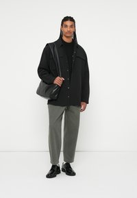 Černá oversized košile, černý svetr, olivově zelené kalhoty a černé šněrovací boty. Modelka nese černou koženou kabelku přes rameno.