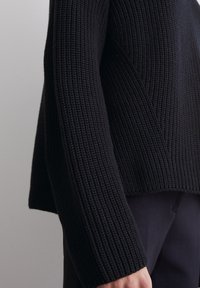 Schwarzer gestrickter Pullover mit einem strukturierten Rippenmuster, der einen verkürzten Saum und weite Ärmel aufweist. Das Material wirkt dick und gemütlich.