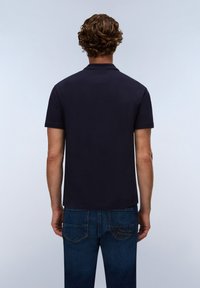 Hombre de pelo rizado visto desde atrás con un polo liso de manga corta azul marino oscuro y vaqueros azules sobre un fondo neutro.