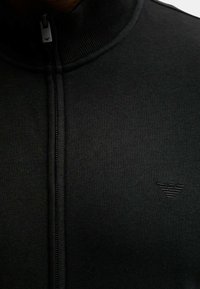 Svart zip-up jacka med hög krage, tillverkad av stickat material. Har en diskret präglad logotyp på bröstet och en texturerad yta.
