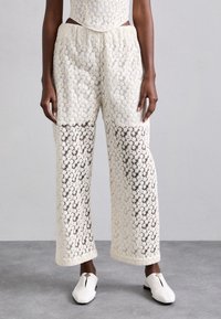 Hvite blondebukser med blomstermønster, semi-gjennomsiktige seksjoner, elastisk midje, og vide ben. Brukt med hvite slip-on sko.
