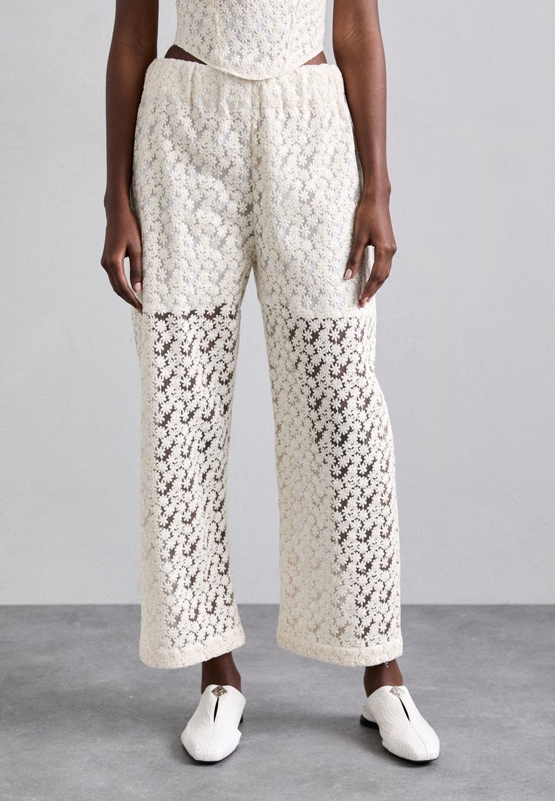 Hvite blondebukser med blomstermønster, semi-gjennomsiktige seksjoner, elastisk midje, og vide ben. Brukt med hvite slip-on sko.
