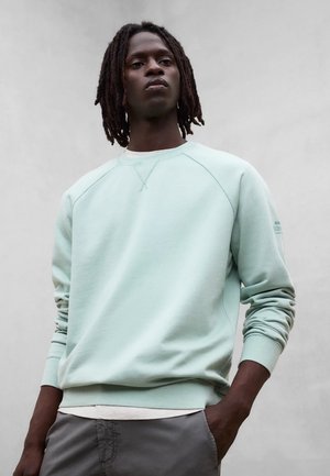 Uomo con dreadlocks indossa una felpa verde chiaro e pantaloni grigi, in piedi con le mani in tasca davanti a uno sfondo grigio uniforme.