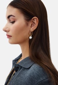 Øreringe i guldfarve med kædedesign og en fremtrædende stor hvid perle. Modellen bærer en mørkegrå top med krave og en guldkæde.