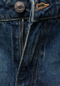Jean en denim bleu partiellement déboutonné avec une fermeture éclair en métal visible, des surpiqûres en cuivre et un bouton métallique portant l'inscription "King Jeans Established 1984".