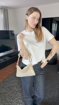 Mala de malha dourada com design curvo, alça fina, combinada com uma t-shirt branca e jeans escuros. O fundo apresenta um móvel para TV escuro.