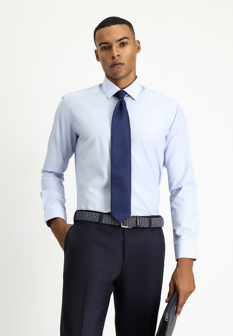 Camicia azzurro chiaro con colletto a punta, abbinata a una cravatta blu navy. Pantaloni scuri su misura e una cintura con motivo. Tieni in mano una pochette scura.