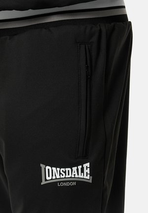 Pantaloni atletici negri au o textură netedă, o buzunar cu fermoar și un brâu cu dungi gri, având un logo alb și argintiu pe coapsă.