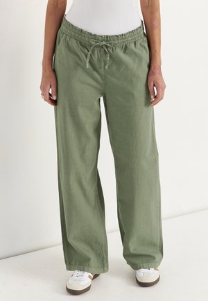 Persoon die losse olijfgroene broek met trekkoord, witte sneakers en een strak wit topje draagt, staand tegen een effen achtergrond.