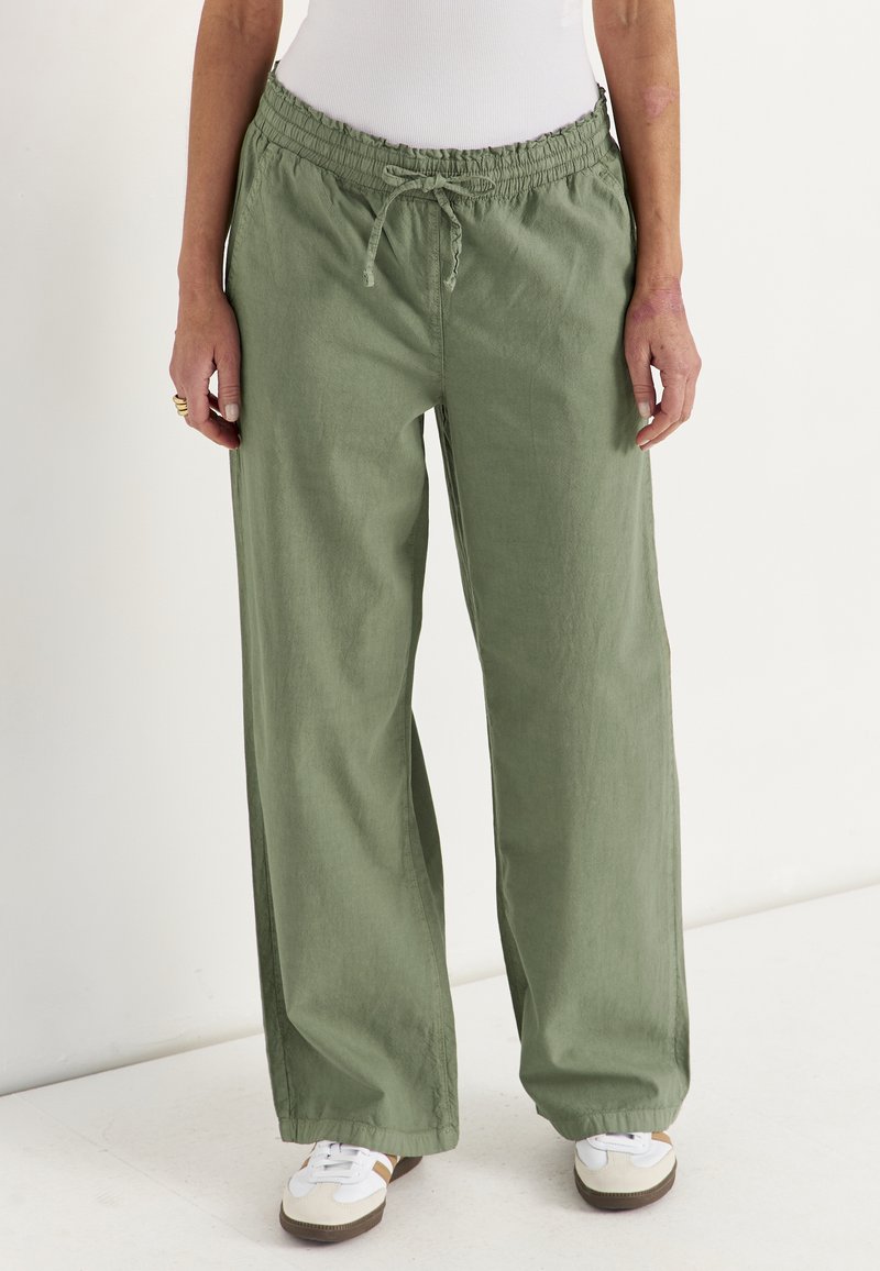 Person trägt lockere olivgrüne Hose mit Kordelzug, weiße Sneaker und ein weißes, eng anliegendes Oberteil, steht vor einem schlichten Hintergrund.