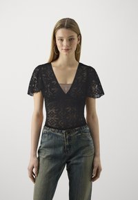 Schwarzer Spitzen-Bodysuit mit tiefem V-Ausschnitt und kurzen, schwingenden Ärmeln, verziert mit Diamantmustern. Kombiniert mit hochgeschnittenen blauen Jeans.