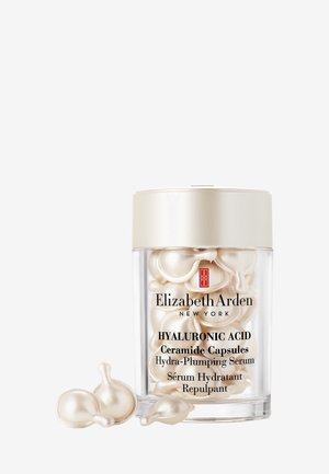 Conteneur cylindrique transparent avec un couvercle doré, contenant des capsules d'acide hyaluronique de couleur crème. Les étiquettes en noir et rouge détaillent le nom du produit et son utilisation.
