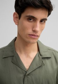 Chemise à manches courtes couleur vert olive en tissu léger, dotée d'un col à revers et d'une fermeture par bouton, avec une texture lisse et un design minimaliste.