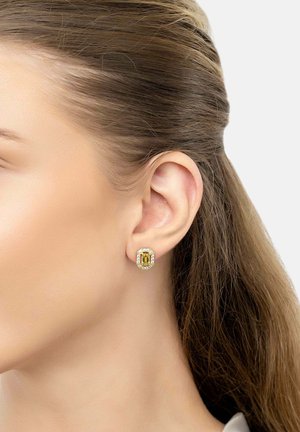 Oreja de mujer con un pendiente rectangular de piedra preciosa amarilla rodeado de pequeñas piedras transparentes, con cabello castaño claro recogido.