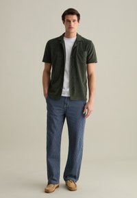 Korte mouwen donkergroen overhemd met een zak, gecombineerd met losse navy en witte gestreepte broek. Het model draagt beige instappers.