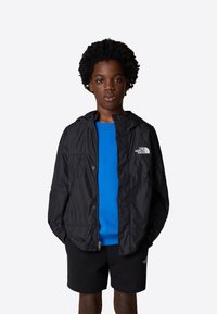 Svart lättjacka med huva, med tryckknappar och en logotyp på vänster bröst, bärs över en blå tröja och svarta shorts.