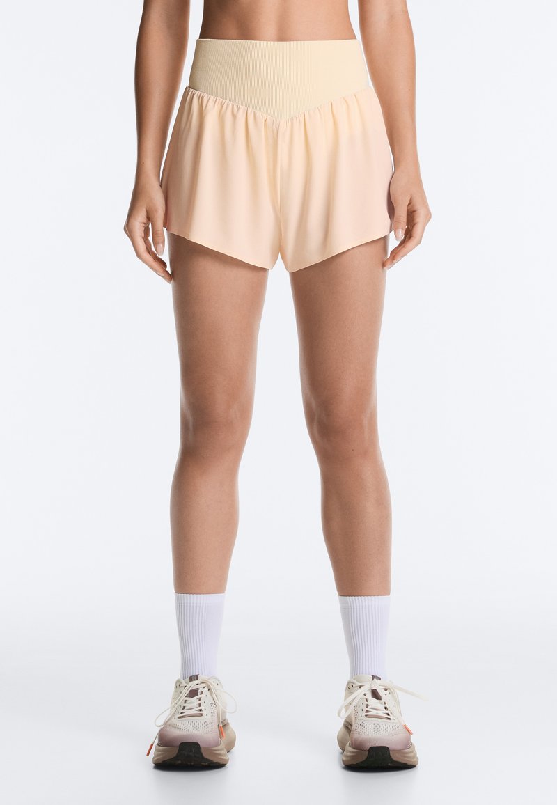 Femme portant un short de sport taille haute couleur pêche clair, des chaussettes blanches classiques et des chaussures de course beige, debout sur fond blanc.