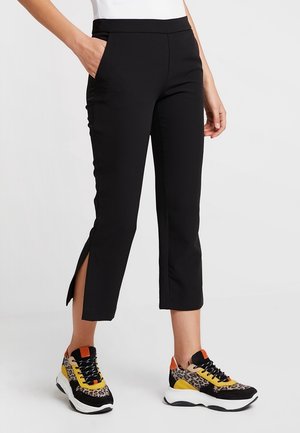 Vrouw die zwarte cropped broek met zijsplitten draagt en meerkleurige sneakers met luipaardprint met gele en oranje accenten.