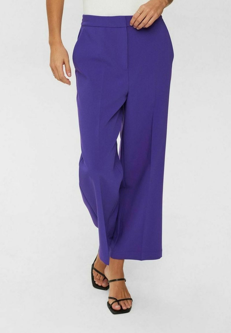 Femme portant un pantalon violet taille haute à jambes larges et des sandales noires à talons avec brides, debout devant un fond uni.