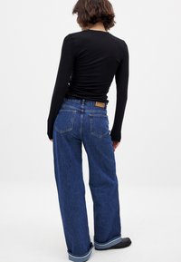Svart långärmad topp i kombination med blå vida denimjeans med en synlig midjebandsetikett och uppvikta ärmslut, presenterad från baksidan.