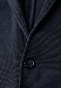 Mörk marinblå jacka med en texturerad tyg, som har breda slag, en svart knapp och fina sömdetaljer.