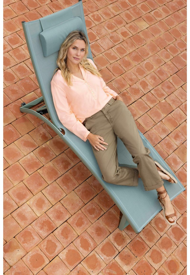 Sedia da lounge reclinabile di colore blu chiaro con poggiatesta, posata su piastrelle in terracotta. Una persona si rilassa in modo informale, indossando una maglietta pesco e pantaloni khaki.