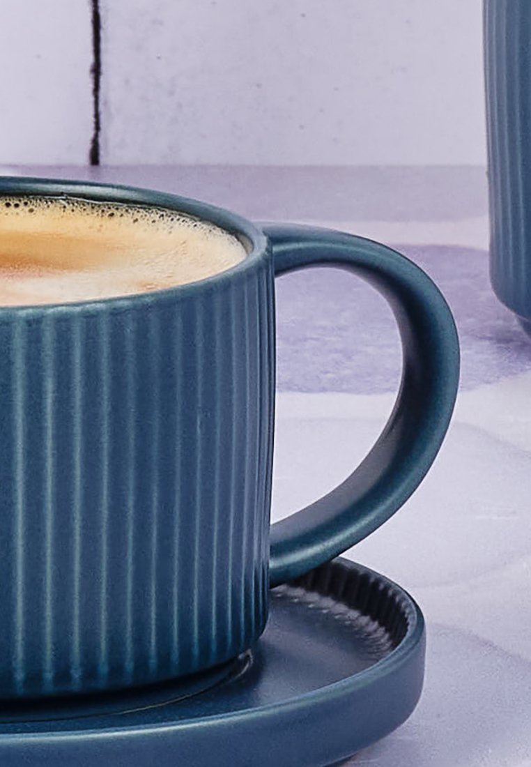Tazza in ceramica blu con texture a righe verticali, base rotonda e piattino abbinato, riempita di caffè, con una superficie schiumosa.