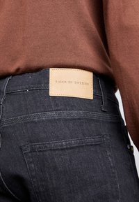 Svarta jeans i denim med en läderetikett som säger "TIGER OF SWEDEN." Tyget har en slät yta och visar detaljer i bakfickornas sömmar.