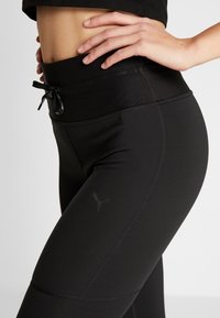 Leggings sportivi neri realizzati in tessuto liscio, con un alto elastico in vita dotato di cordino, cuciture laterali e un piccolo logo vicino alla coscia.
