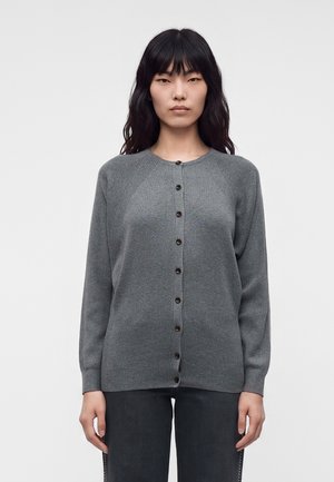 Kvinde med langt sort hår iført en grå cardigan med knapper og mørke bukser, stående mod en ensfarvet lysegrå baggrund.