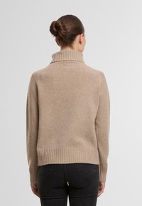 Beige gestrickter Pullover mit hohem Kragen, gerippten Bündchen und Saum. Weiche Textur und lockere Passform, mit einer Rückennaht-Detail.