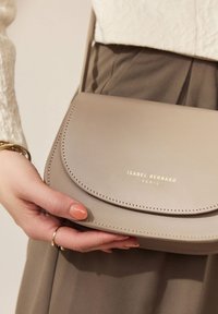Beige Lederhandtasche mit einem gebogenen Klappdeckel, goldfarbener Prägung "ISABEL BERNARD PARIS" und sichtbaren Nähdetails, gehalten in einer Hand.