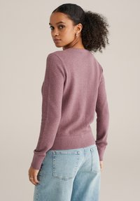 Gebreide trui in stoffig roze, met een getextureerd patroon, lange mouwen en een geribde zoom. Draagt een lichtblauwe, hooggewate jeans.