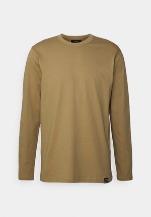 Prosty jasnobrązowy longsleeve z okrągłym dekoltem i małą czarno-białą metką w pobliżu dołu na neutralnym tle.