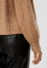 Hellbraunes, gestricktes Sweatshirt mit lockerer Passform und gerippter Struktur, kombiniert mit schwarzen Lederhosen. Das Sweatshirt hat breite, weiche Bündchen.