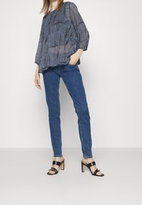 Flowy, sheer navy blouse with wave patterns; long sleeves; paired with dark blue jeans; black block heel sandals with two straps.