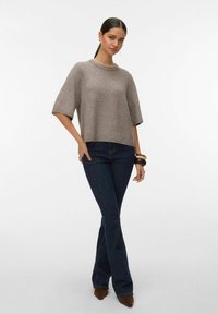 Femme aux cheveux foncés attachés en queue de cheval basse, portant un pull gris à manches courtes, un jean flare bleu foncé, des chaussures marron pointues et des bracelets.