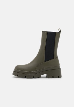 Støvle med chunky sål i olivengrøn og sort elastisk sidepanel samt trækflig på ensartet lys baggrund.