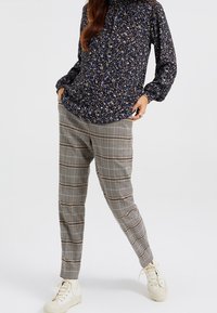 Kvinna i marinblå blommig långärmad blus, beige rutiga tapered byxor och vita högskurna sneakers, står med händerna i fickorna.