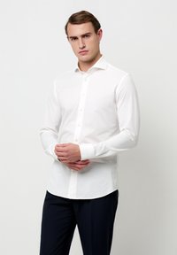 Camicia bianca a maniche lunghe con colletto classico, caratterizzata da una chiusura frontale con bottoni e una leggera lucentezza, abbinata a pantaloni scuri. Design semplice e aderente.