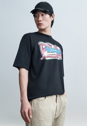 Νεαρός άνδρας με μαύρο καπέλο, μαύρο T-shirt με πολύχρωμο σχέδιο, και ανοιχτό μπεζ παντελόνι, στέκεται μπροστά σε απλό γκρι φόντο.