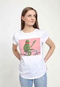 Bílá tričko s potiskem zelené žáby na modrém bicyklu na růžovém pozadí, krátké rukávy a přiléhavý střih.