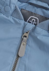 Ljusblå jacka i tyg med en slät yta, med en silverfärgad dragkedja och ribbade kragar. Inkluderar en etikett med en logotyp.