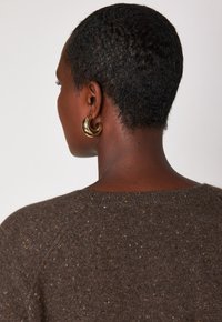 Person mit kurzem, strukturiertem Haar, die große goldene Creolen trägt und einen braun gesprenkelten Strickpullover anhat, von hinten vor einem schlichten Hintergrund gezeigt.